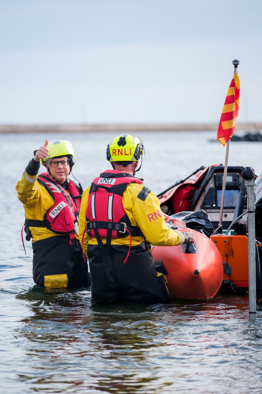 An RNLI crew member stood next to D-Class Lifeboat in knee deep water preparing to launch, showing a thumbs up to the shore crew.