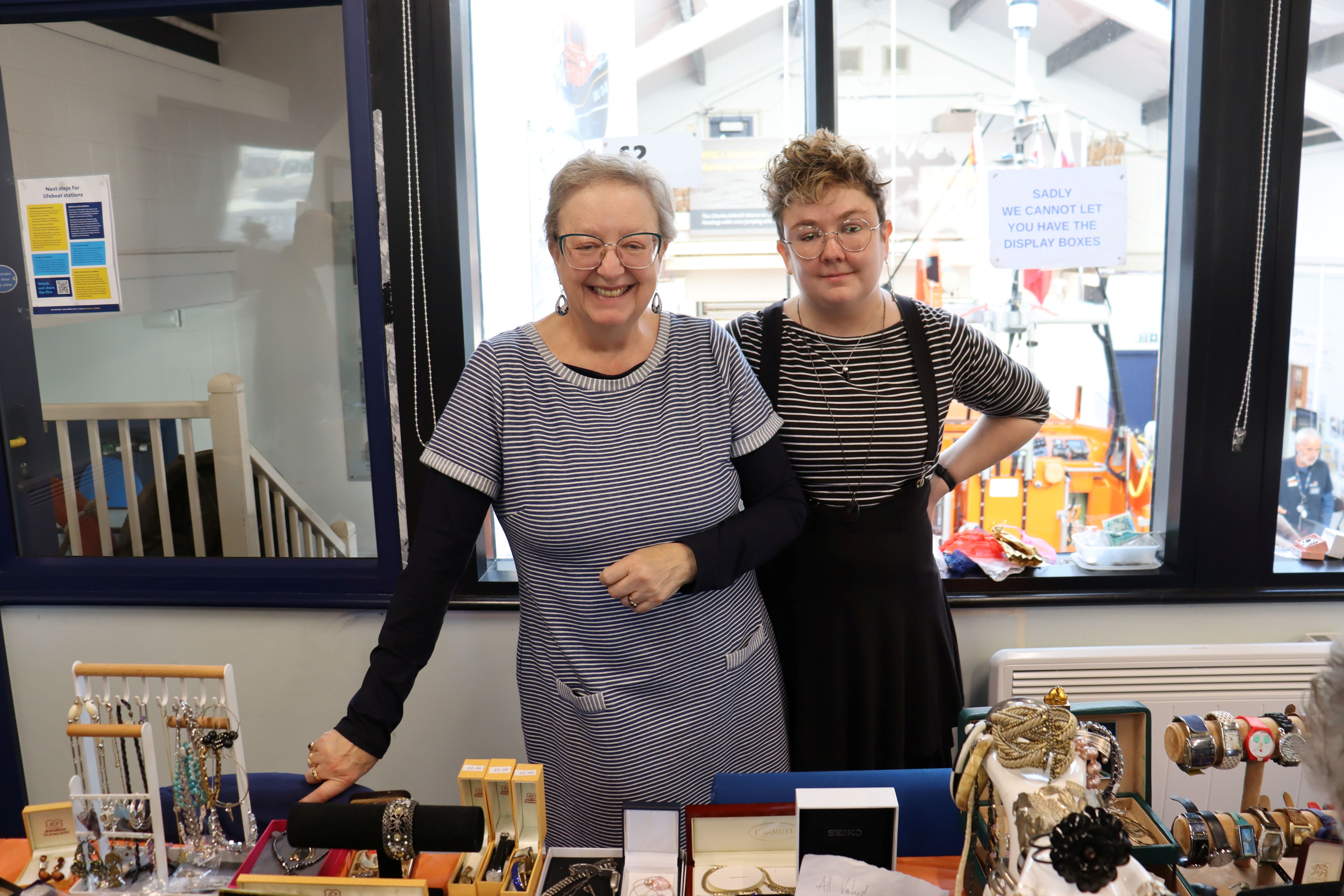 Two volunteers at the sale, with jewellery in front of them and a lifeboat as a backdrop