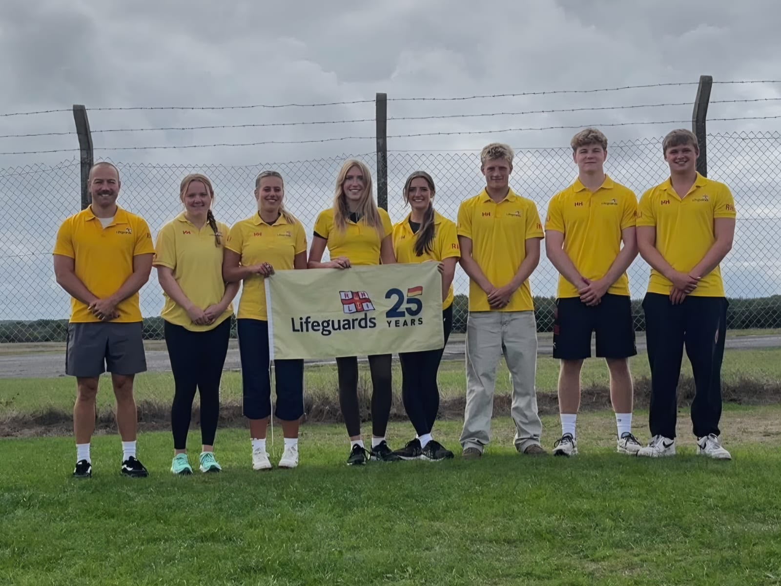 Landscape image of lifeguards lined up with RNLI lifeguard 25th anniversary flag