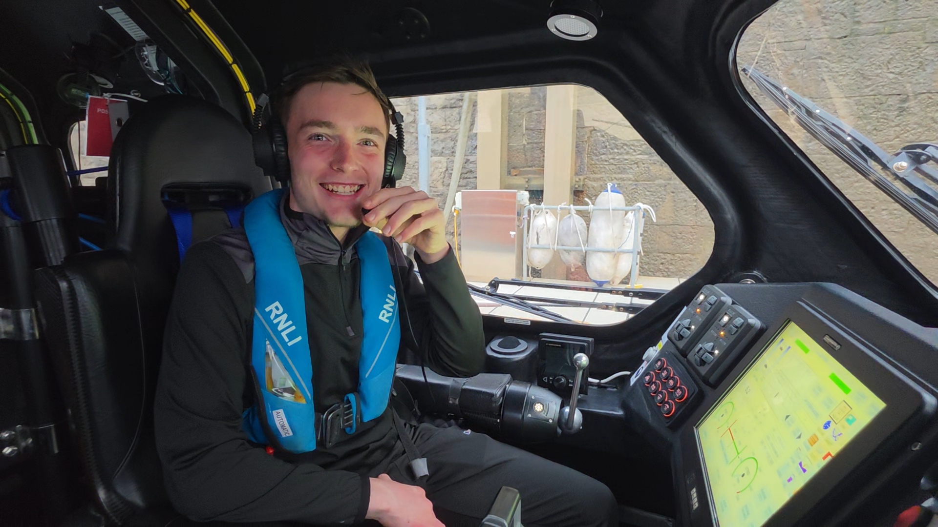 A young man in the seat of a lifeboat with a headset on, smiling.
