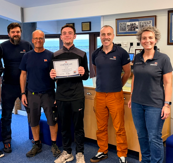 A young lad holding a framed certificate surrounded by volunteers in RNLI kit