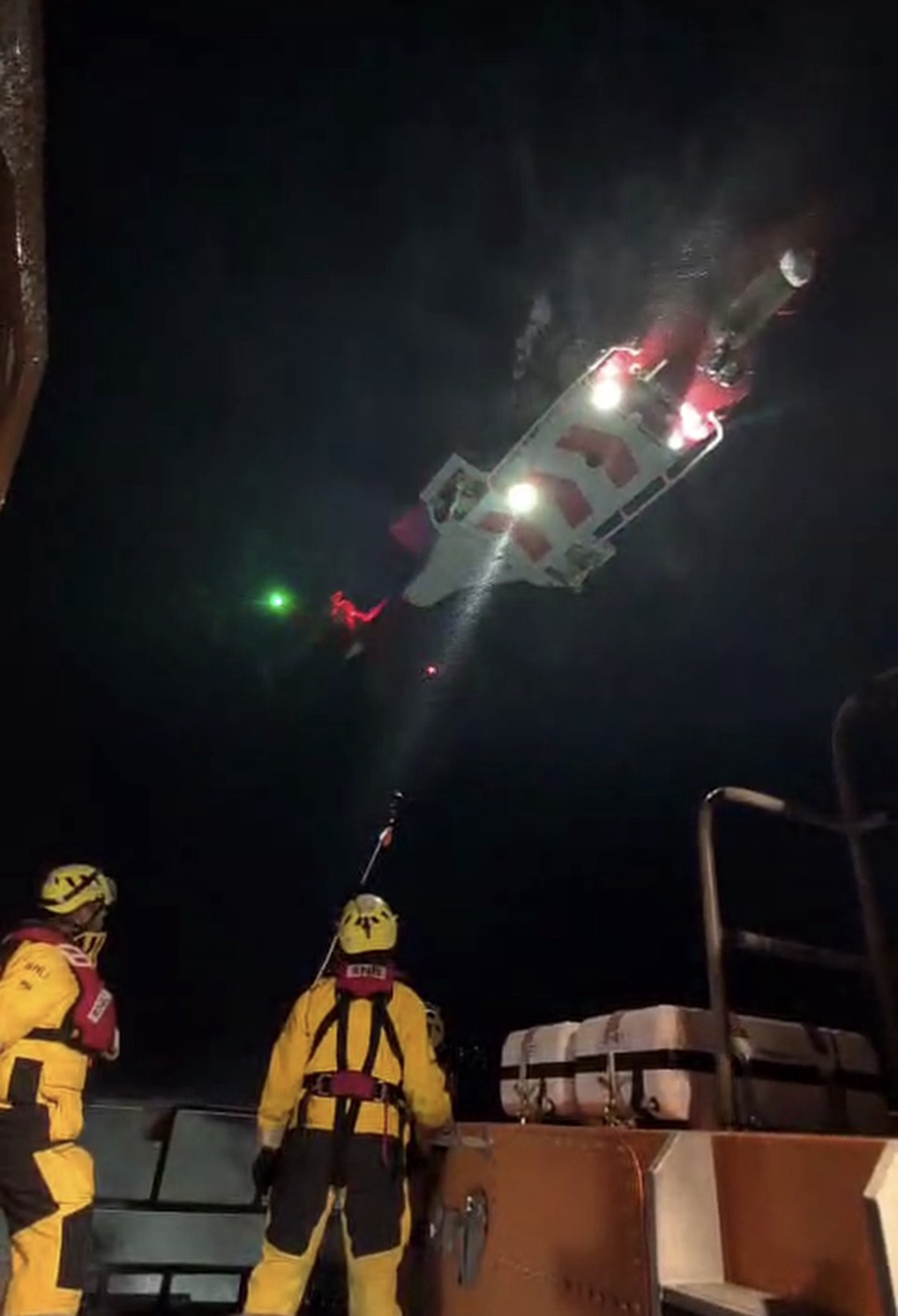 RNLI crew members in yellow protective gear stand on a lifeboat deck at night as a rescue casualty is being lifted by helicopter under bright search lights