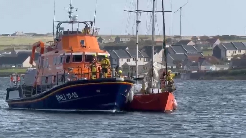 Yacht on an alongside tow with RNLB Margaret Foster as they approach Kirkwall Marina. In the background are houses.