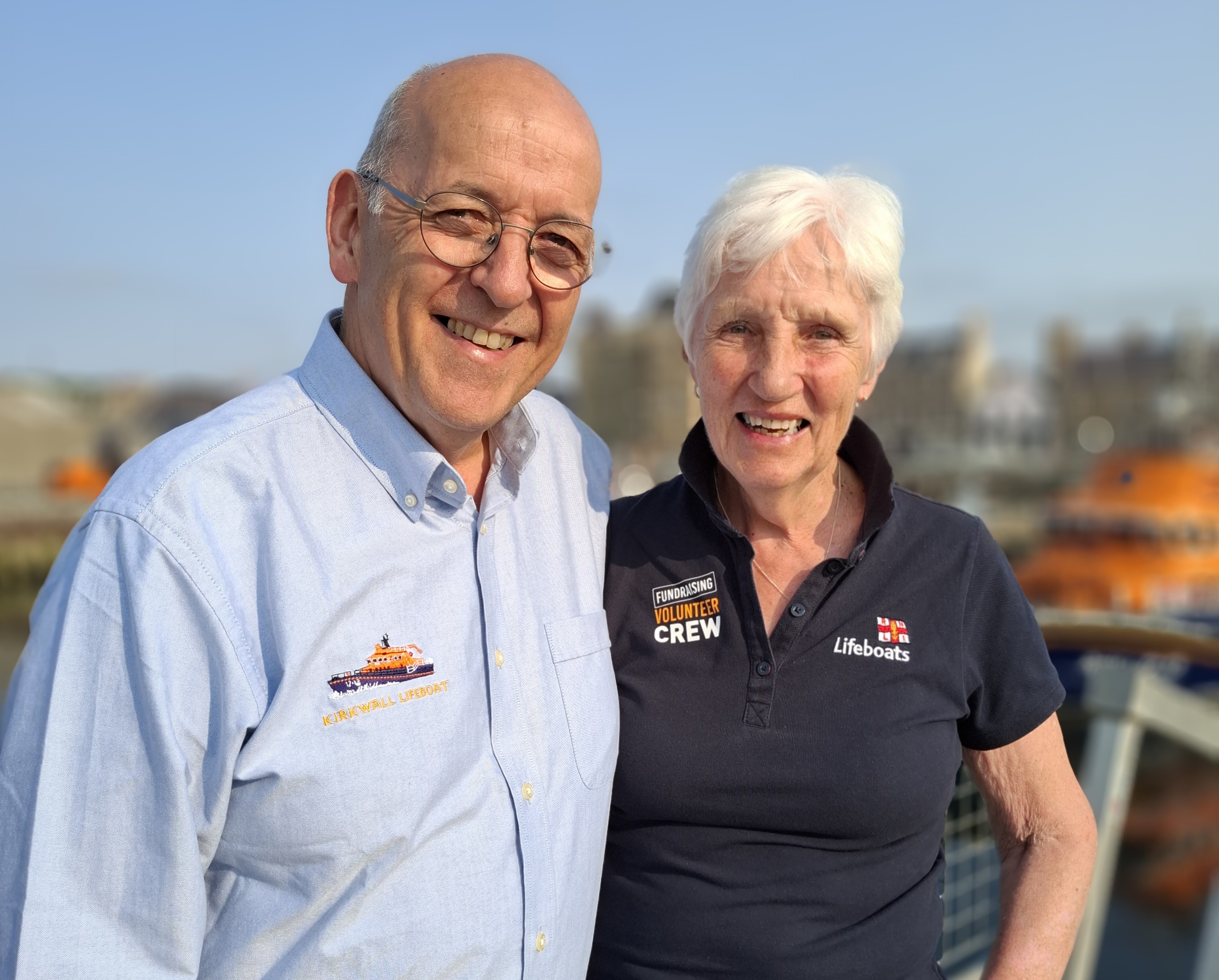 Chris and Moira Irvine, portrait, with lifeboat and pier buildings in the background