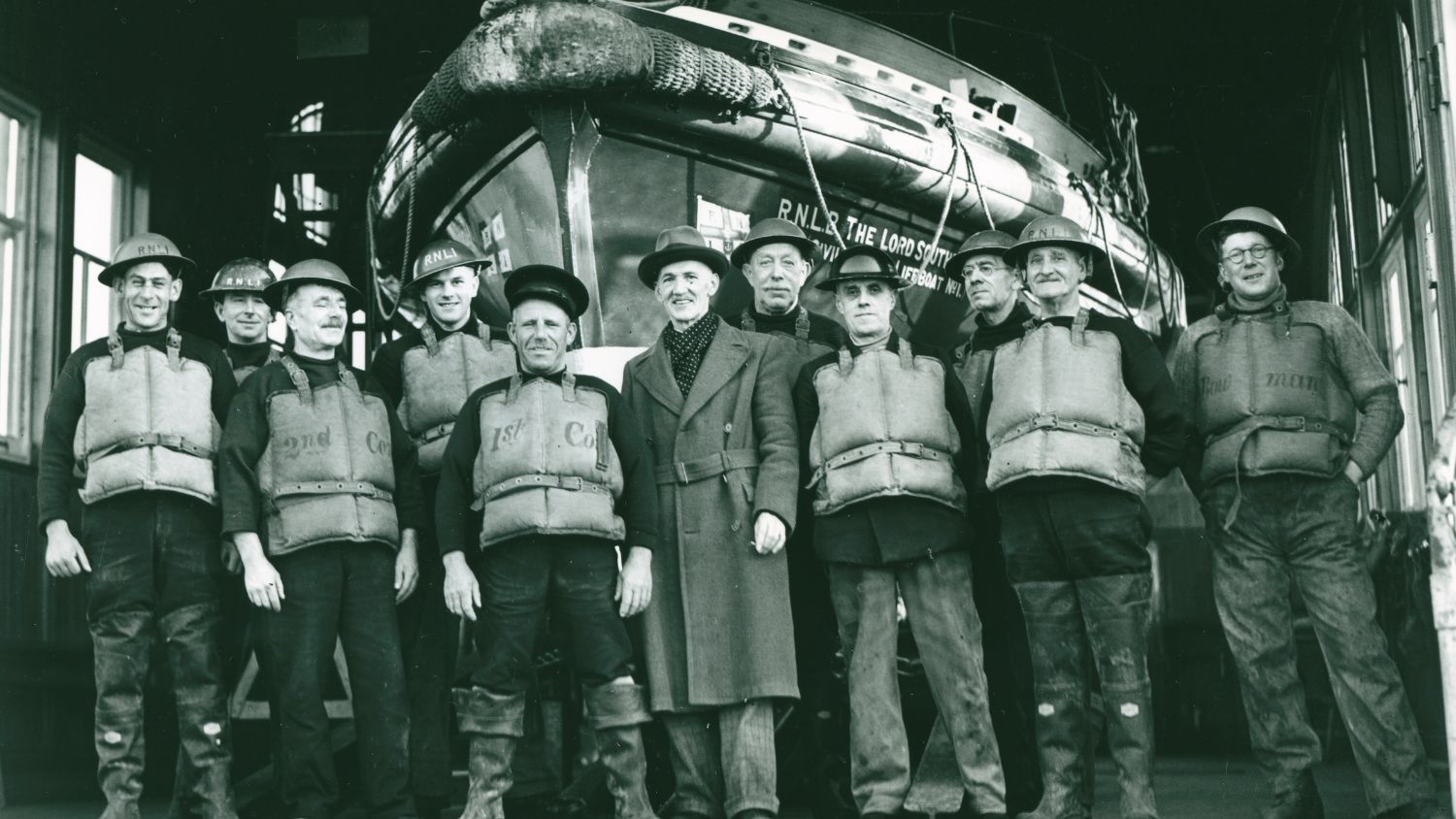 Black and white photo of 11 men stood in a line in front of a lifeboat. 10 of the crew are wearing steel Brodie helmets, many with RNLI printed on the front, and Kapok lifejackets. A man stands in the middle not wearing crew kit. He wears a long coat and smart trousers. They are facing the camera and most are smiling.