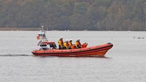 An RNLI lifeboat named "Jean Spier" with five crew members in yellow jackets and helmets travels across calm water, with trees and wildlife visible in the background.