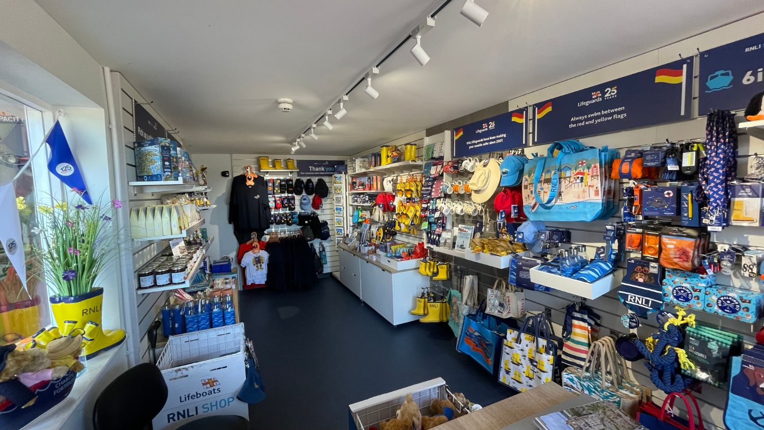 A well-stocked RNLI gift shop displaying a variety of nautical-themed merchandise including clothing, bags, hats, and yellow wellington boots.