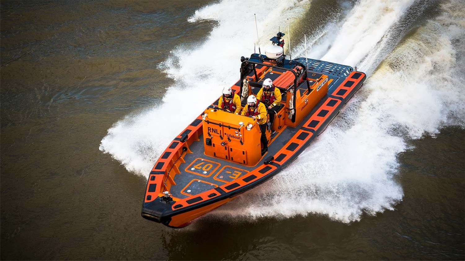 An E class RNLI lifeboat with four crew members in yellow jackets and white helmets speeds through choppy brown water, leaving a large white wake.