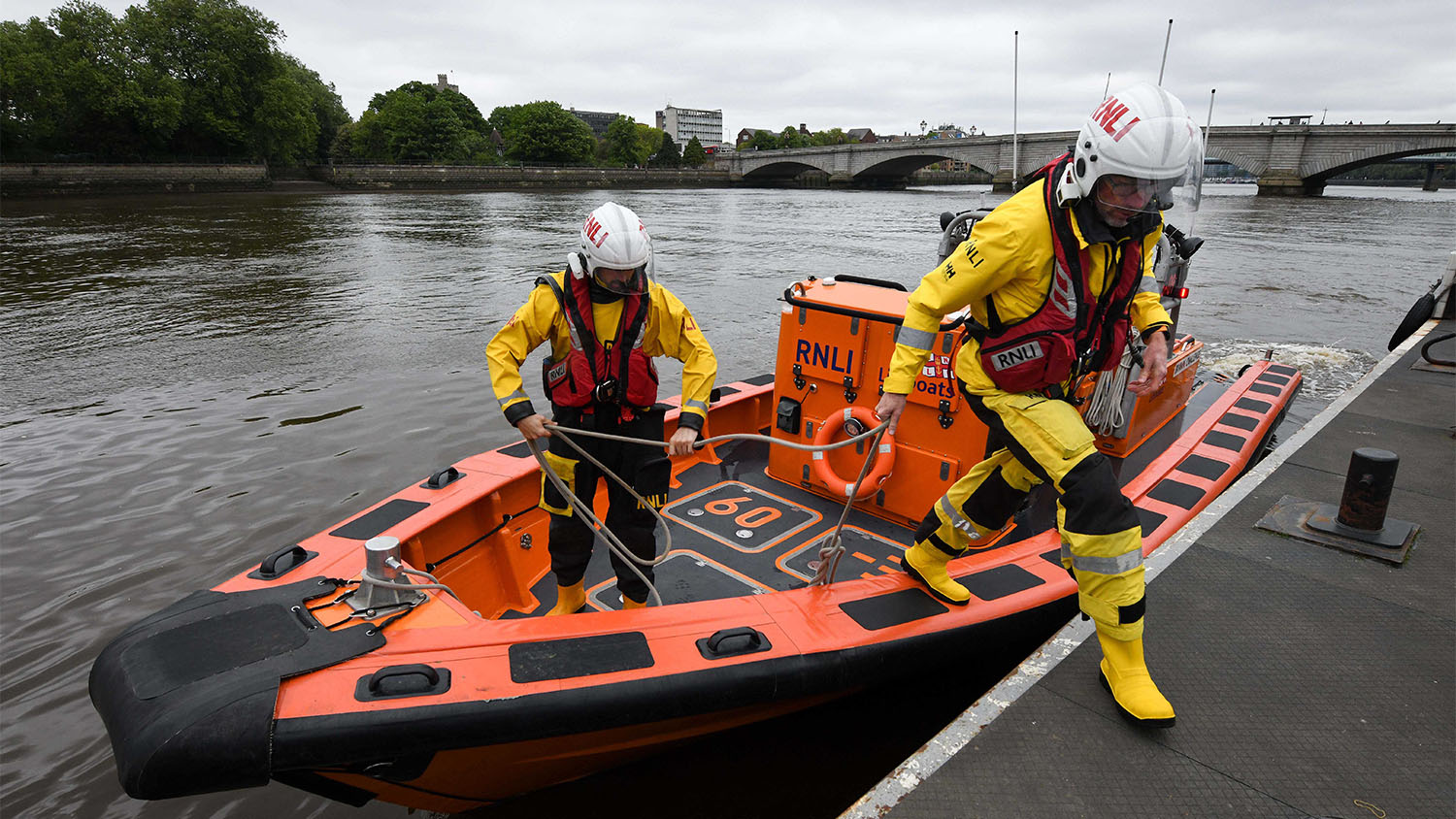 Two RNLI crew members in yellow dry suits and life vests prepare an orange inflatable rescue boat at a dock, with a bridge visible in the background.