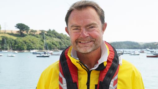 Crew Member Steve Walter smiles as he stands in front of the water. Sailboats can be seen behind him. Credit: RNLI/Nigel Millard