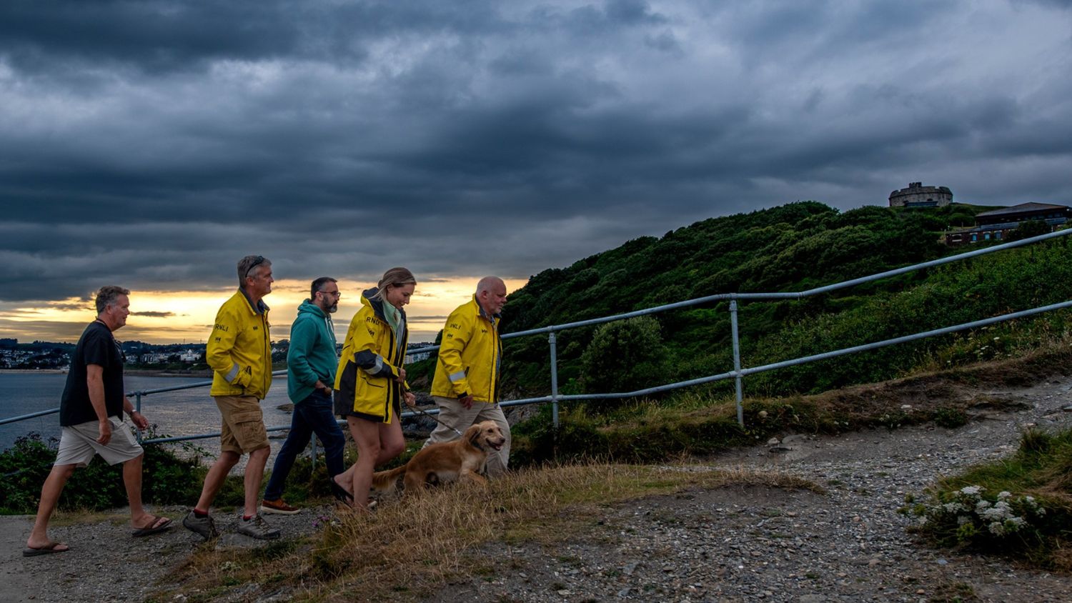 Five people walking along a coastal path at dusk, with three wearing yellow RNLI jackets. A dog walks alongside them. Dark clouds loom overhead, and the sea is visible to the left. Lush green hills and a building sit in the background