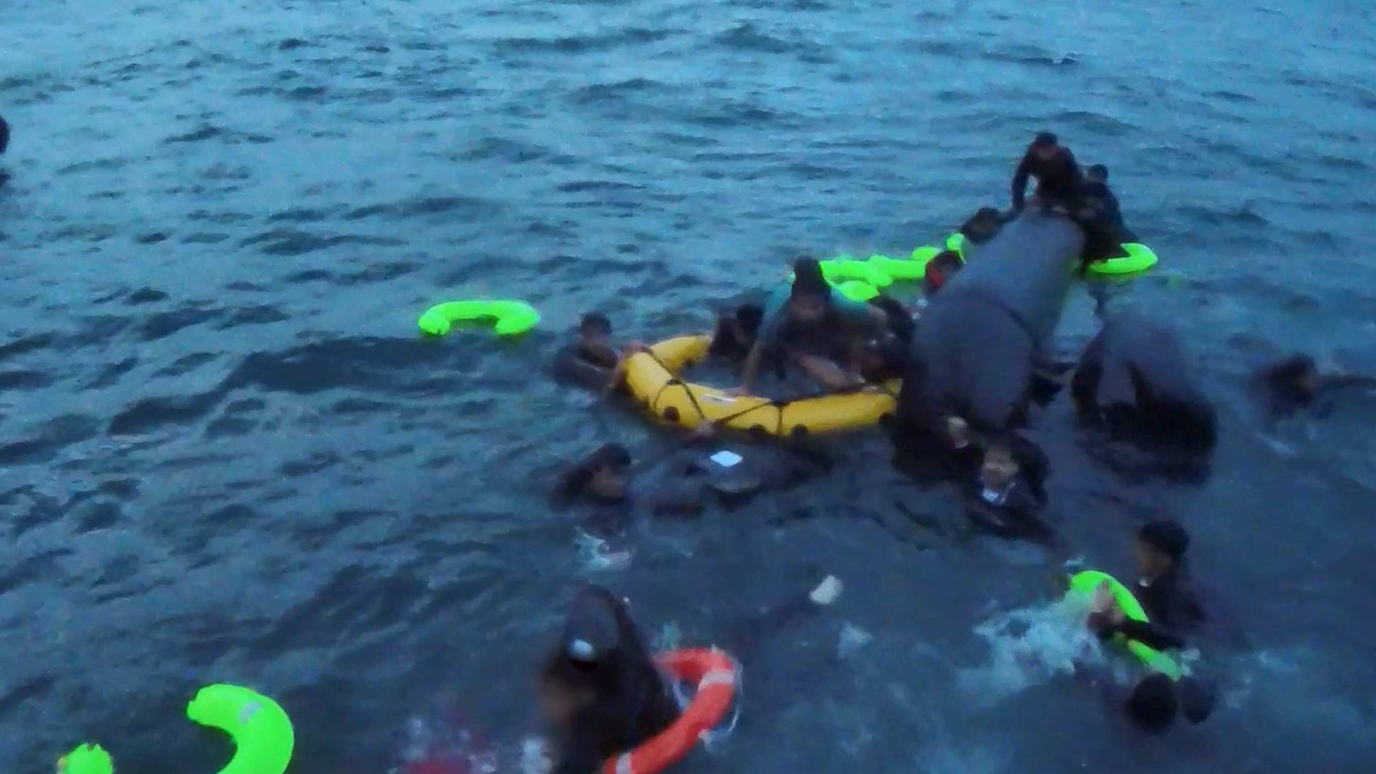 A group of men, women and children float desperately in the water as their boat sinks. Bright green life floats are scattered around them as they each fight for their lives.