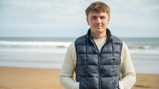 Euan, a man in a white shirt and blue puffer gilet, smiles as he stands on the beach with the water behind him.