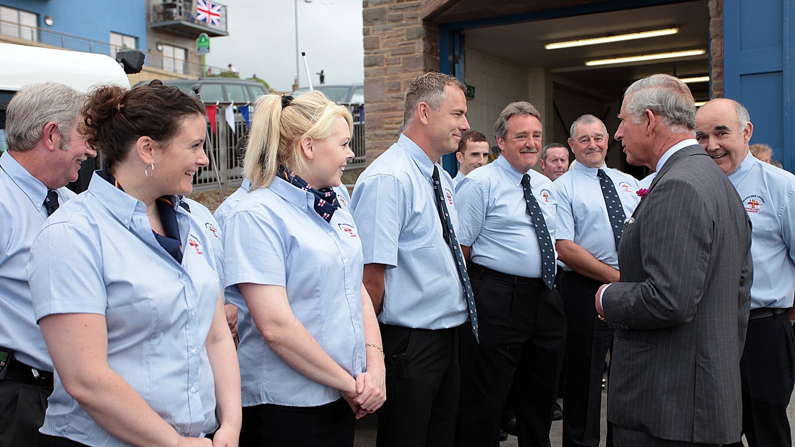 HM King Charles III greets RNLI volunteers wearing blue dress shirts on the slipway outside a lifeboat station