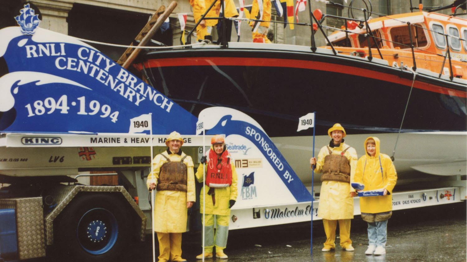 Colour image of four people dressed in historical crew kit with lifeboat behind, taking part in procession
