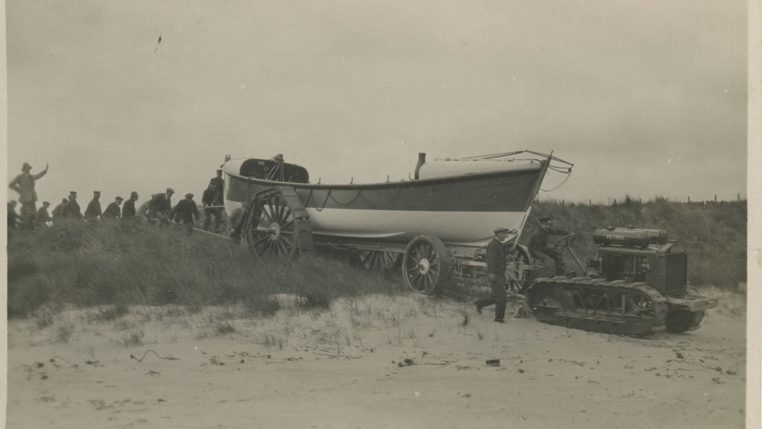 Black and white image of tractor towing lifeboat over sand dune