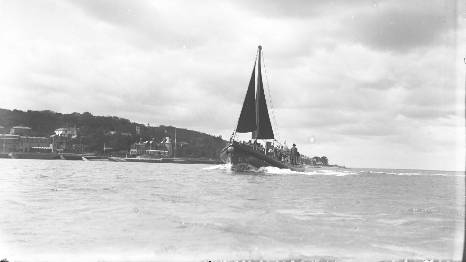 A black and white photo of Holyhead lifeboat, A.E.D., while on trials. The sky is cloudy and coastal land is in the background.