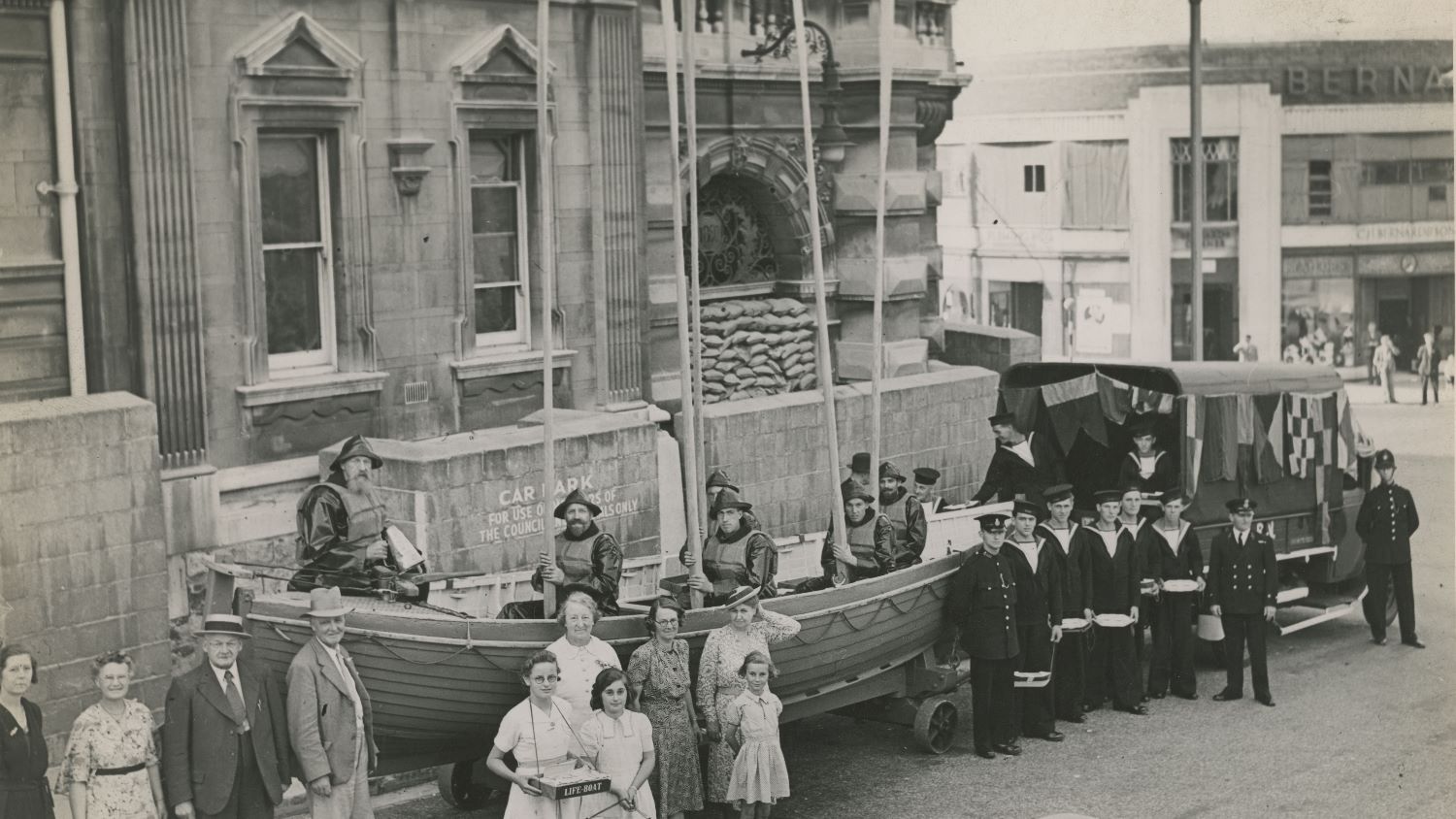 Black and white image of lifeboat pulled by van, lifeboat crew inside with oars raised and fundraisers alongside