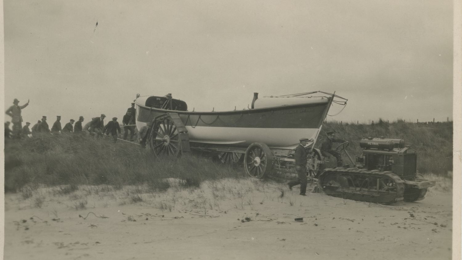 Black and white image of tractor towing lifeboat over sand dune