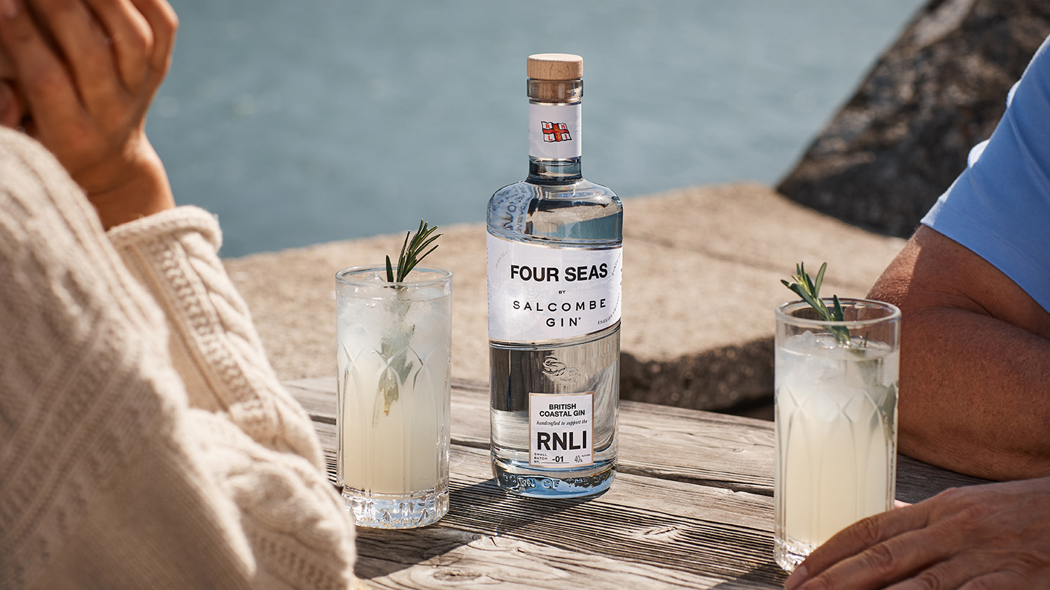 Two people sit at a wooden table, each with a drink and a bottle of Four Seas Salcombe Gin on the table 