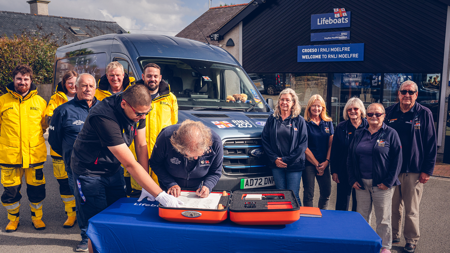 An RNLI volunteer signing the RNLI 200th anniversary scroll on a table in front of RNLI Moelfre Seawatch Centre with other volunteers behind and an RNLI Ford E-Transit van 