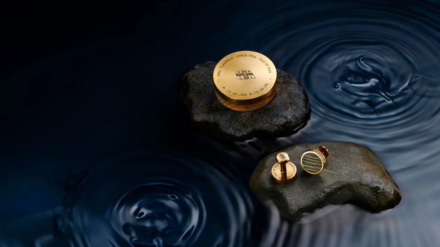 The RNLI 200 Limited Paperweight and RNLI 200 Limited Cufflinks on separate rocks, surrounded by water