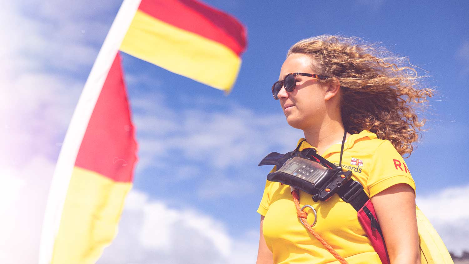 A female lifeguard wearing a yellow lifeguard polo, a radio strapped across her chest, and sunglasses stands staring out to sea beside a red and yellow lifeguard beach flag.