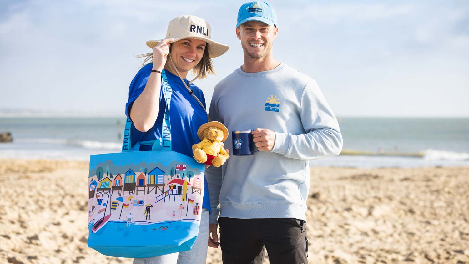 A male and female lifeguard show off the RNLI Shop lifeguard retail range, including a tote bag, teddy bear, mug, hats, t-shirt and sweater
