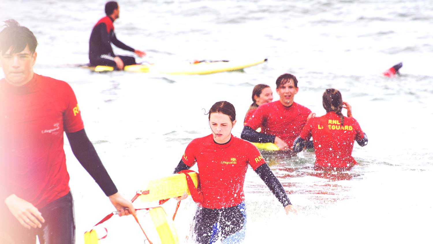 A group of RNLI lifeguards in red t-shirts and black wetsuits train in the surf with rescue tubes 