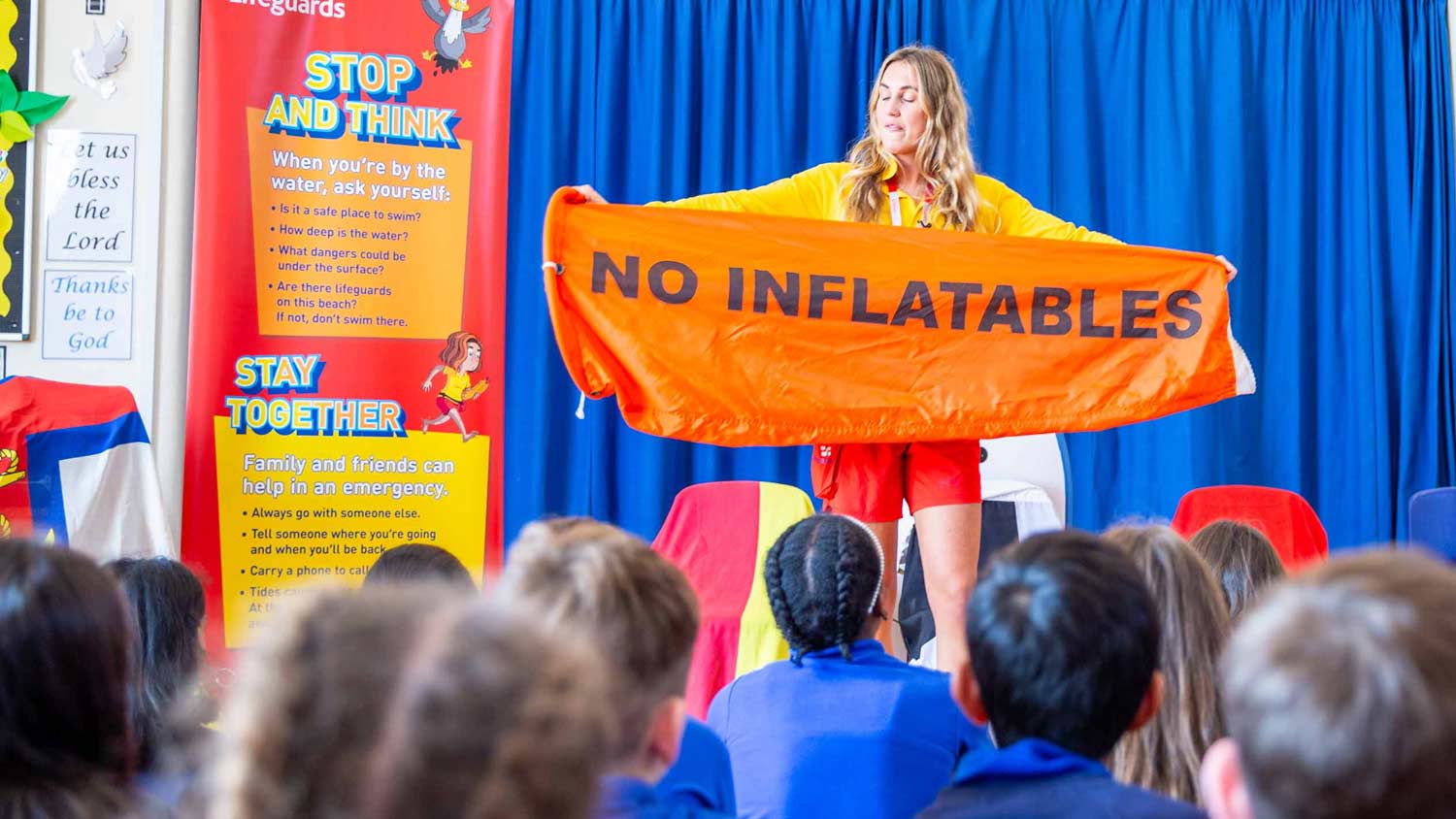 A female RNLI lifeguard holds up an orange flag reading no inflatables to a class of school children inside an assembly hall