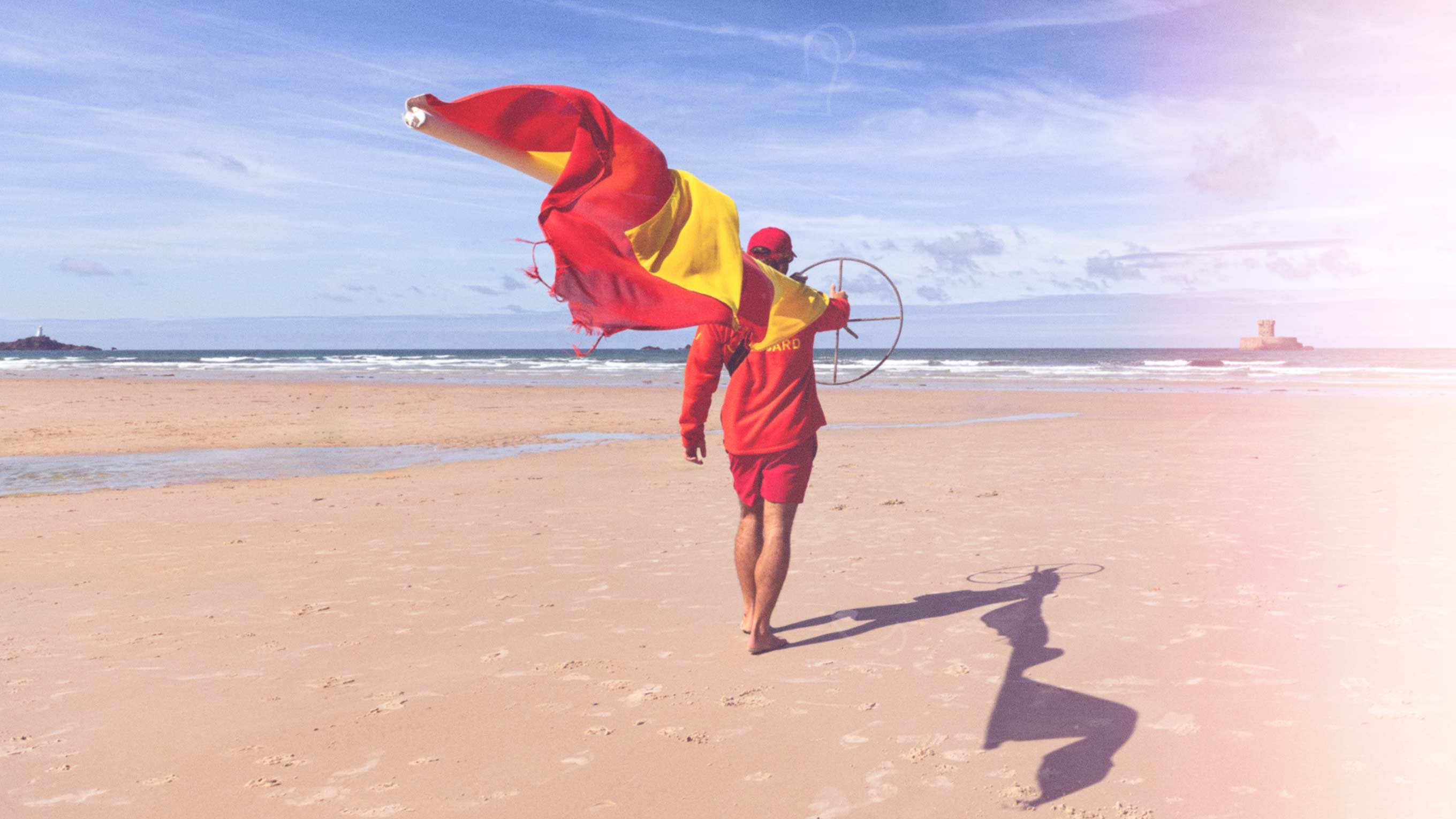 An RNLI lifeguard in red fleece and shorts carries a red and yellow beach flag across the sand to the water's edge. In the distance, St Michaels Mount island can be seen