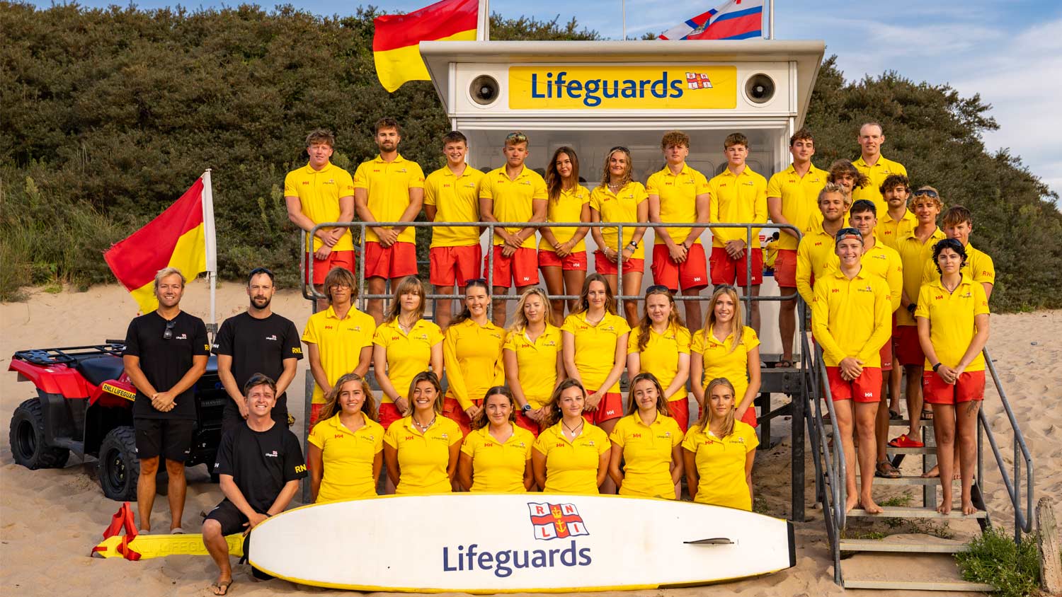 South Pembrokeshire Lifeguard Team pose for a group photo beside a lifeguard hut. Lifeguards wear yellow polo shirts and red shorts, and senior team members are dressed in black