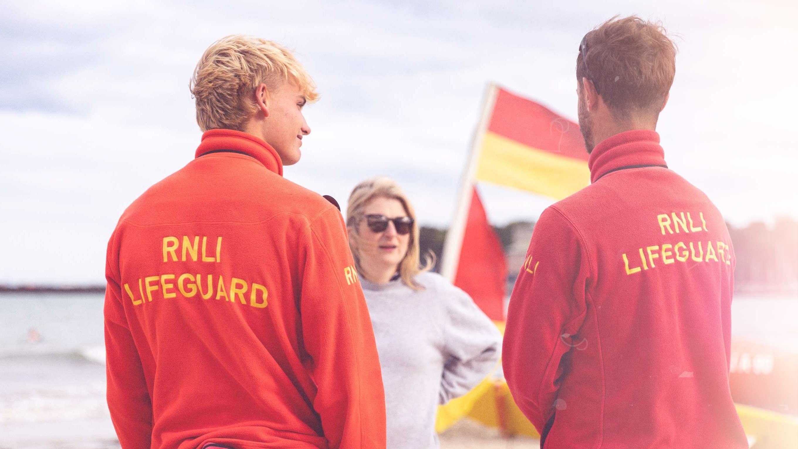 Two lifeguards wearing red RNLI lifeguard fleeces talk with a member of the public on the beach. In the background, a red and yellow beach flag flutters in the wind.