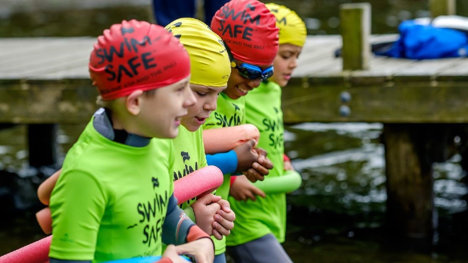 Four children of different ethnicities wade into the water together beside a jetty at a lake, linking arms and holding tube floats under their armpits. They wear red or yellow swim hats, neon green Swim Safe T-shirts and happy smiles on their faces!