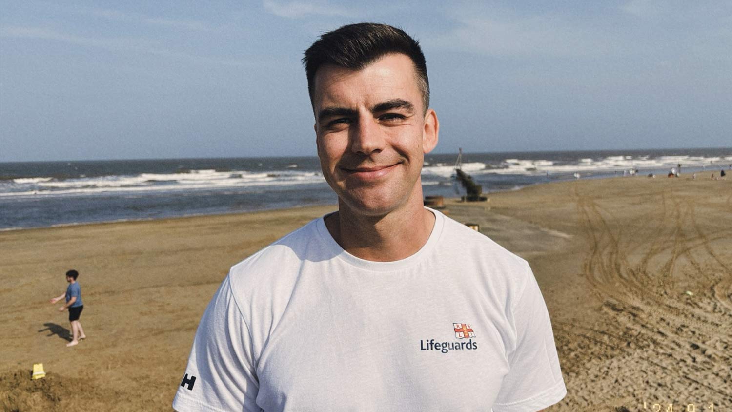 RNLI Lifeguard Supervisor Arun Gray stands on the beach in a white t-shirt