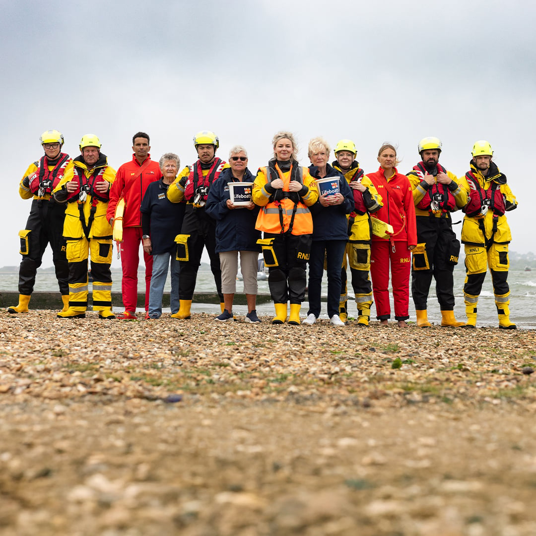 A group of RNLI lifesavers are standing in a line on a pebbly beach, all looking at the camera. Some of them are smiling, and some of them have a neutral expression. There are crew members, lifeguards and fundraising volunteers in the photo. The sea is behind them, and the sky is cloudy.
