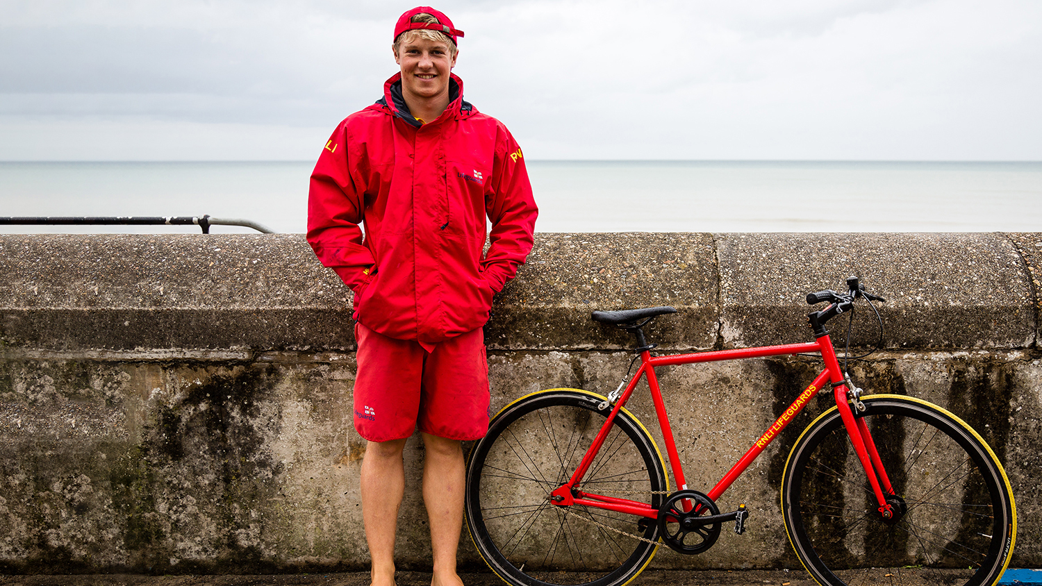 A landscape portrait of a white lifeguard with short blonde hair standing next to a red lifeguard bicycle on a promenade. The lifeguard is wearing lifeguard kit of red shorts, a red waterproof jacket and red baseball cap. The bike is beside them, leaning against a concrete wall with a calm sea in the background and cloudy white sky.