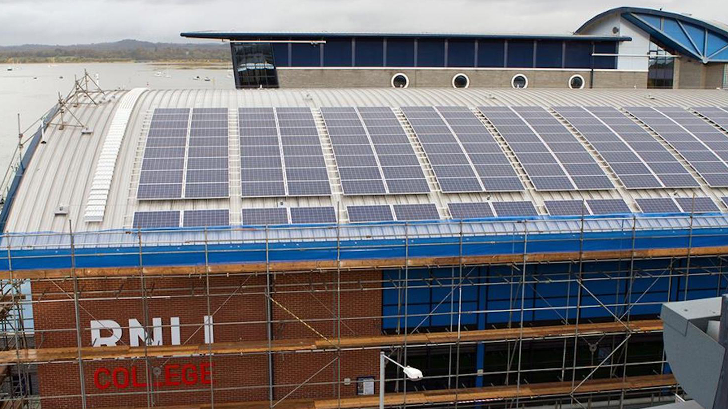 A view of the RNLI’s Sea Survival Centre main building from an elevated aspect, showing the curved roof covered in multiple solar photovoltaic (PV) panels. The building is surrounded by scaffolding and the RNLI Support Centre office building can be seen in the background.
