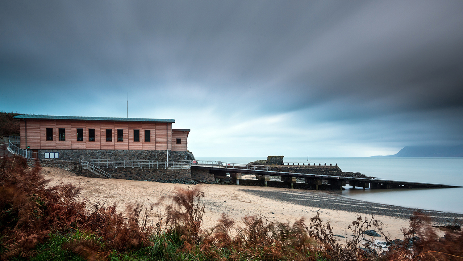 An autumnal view along the coast towards Porthdinllaen Lifeboat Station on the Llyn Peninsula. The lifeboat station is clad in wood and designed for reduced environmental impact. In the foreground is a coastal path with grasses and other wild plants, the station is situated on a yellow sandy beach, and the sea is calm, with mountains visible in the background, beyond the water.