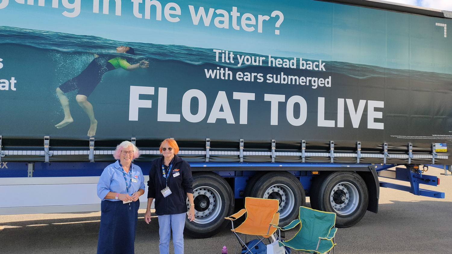 Two RNLI volunteers are standing in front of the Scania trailer, which is wrapped in the RNLI’s Float to Live campaign ad. They are both smiling at the camera. There are two camping chairs beside them, along with some Float to Live flyers and their personal belongings.
