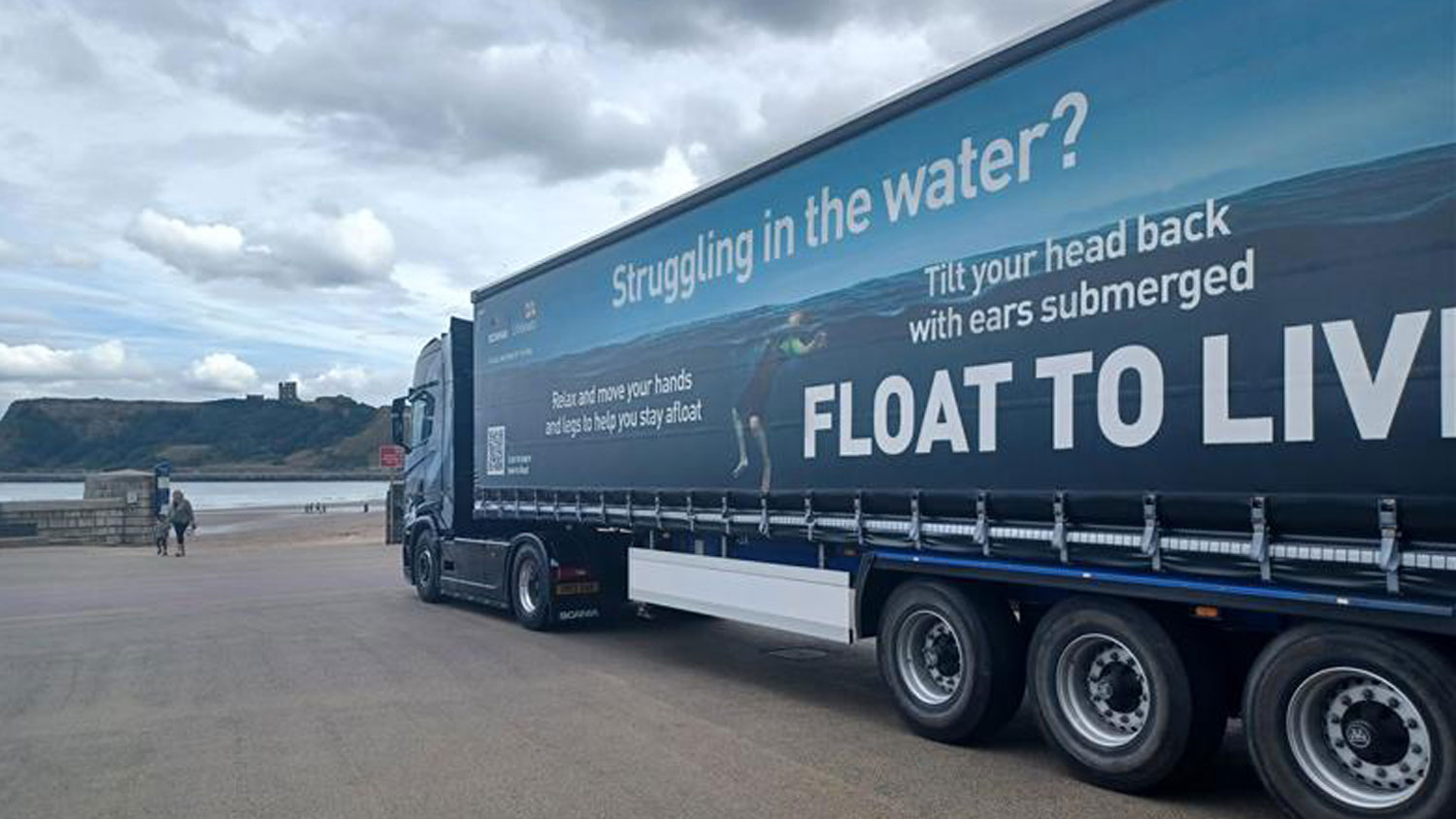 A Scania trailer is parked near the Scarborough coastline. The trailer is wrapped in the RNLI’s Float to Live campaign ad. The sky is very cloudy.