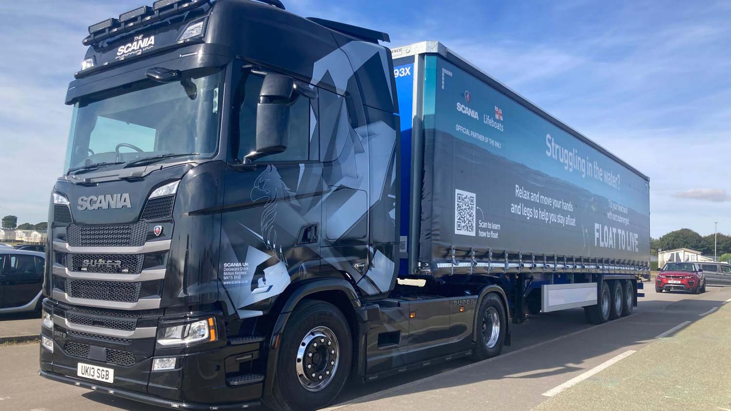 A Scania trailer is parked on the side of a road in Bridlington. The trailer is wrapped in the RNLI’s Float to Live campaign ad. There is a red car parked behind the trailer. The sky is blue with very few clouds.