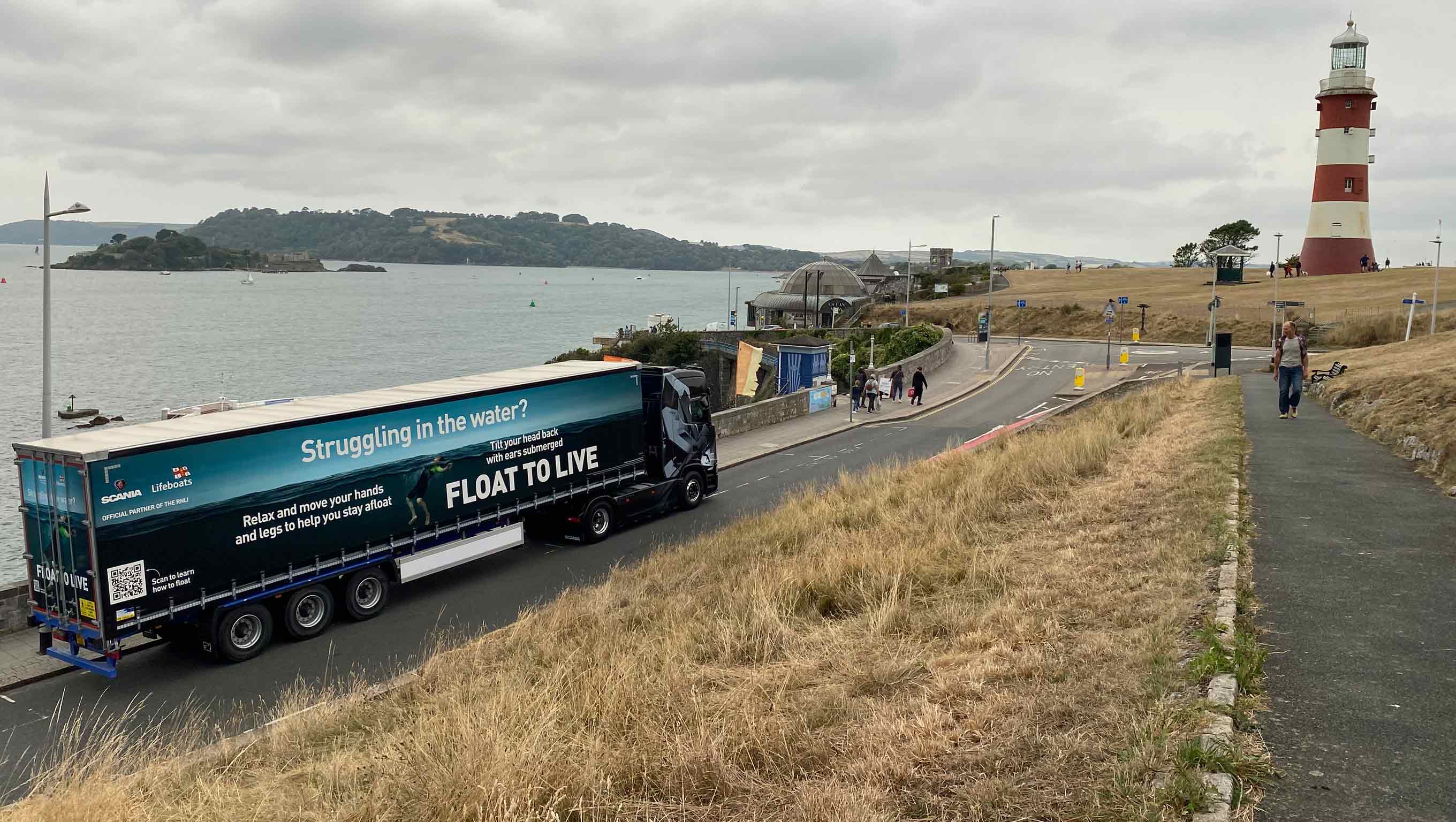A Scania trailer is parked on the side of a road along the Plymouth coastline. The trailer is wrapped in the RNLI’s Float to Live campaign ad. The lighthouse at Plymouth Hoe is in the distance. The sky is cloudy.