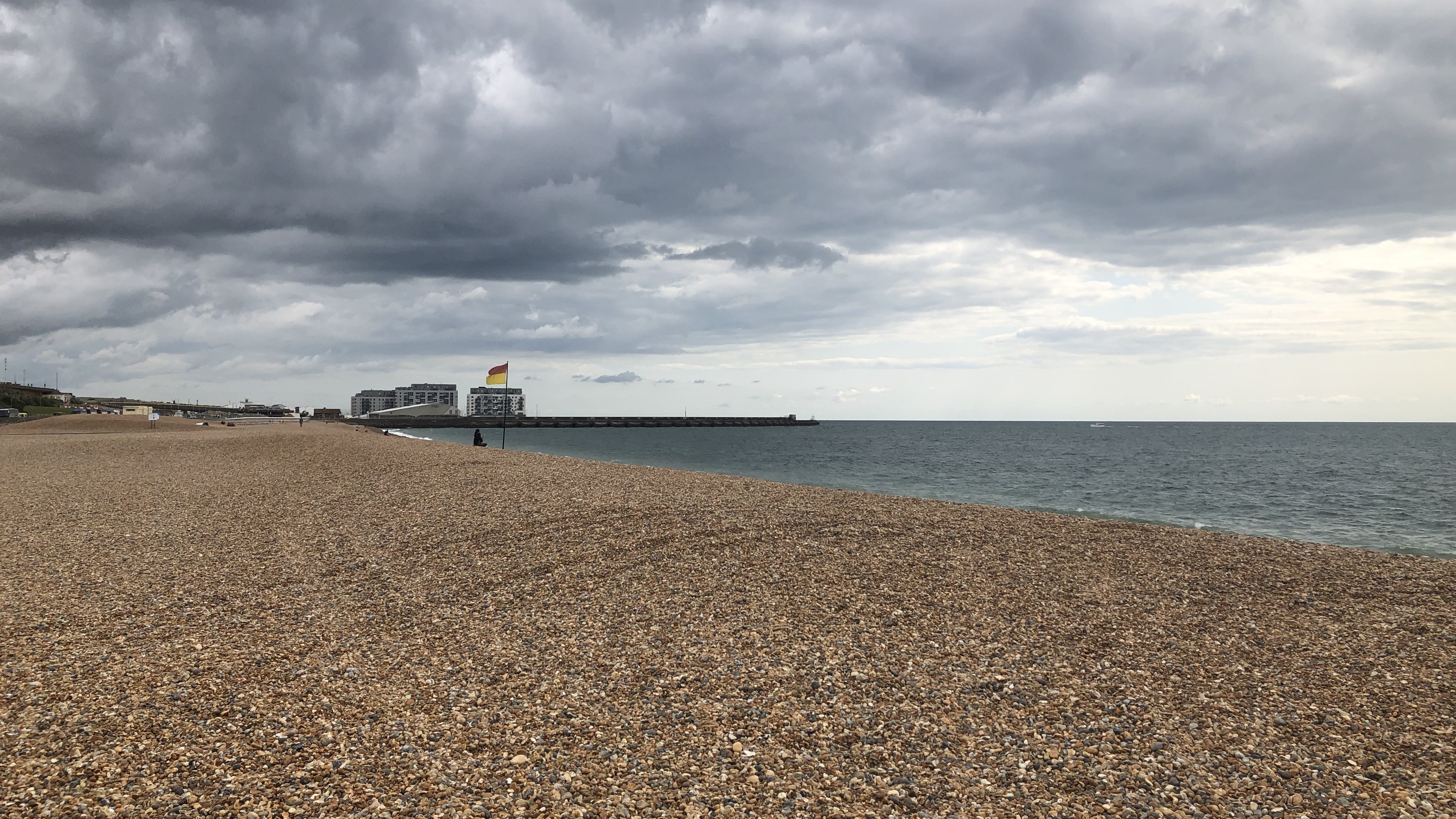 A landscape image of Dukes Mound beach on an overcast day. The beach is a pebble beach. In the distance, a pier can be seen over the sea.