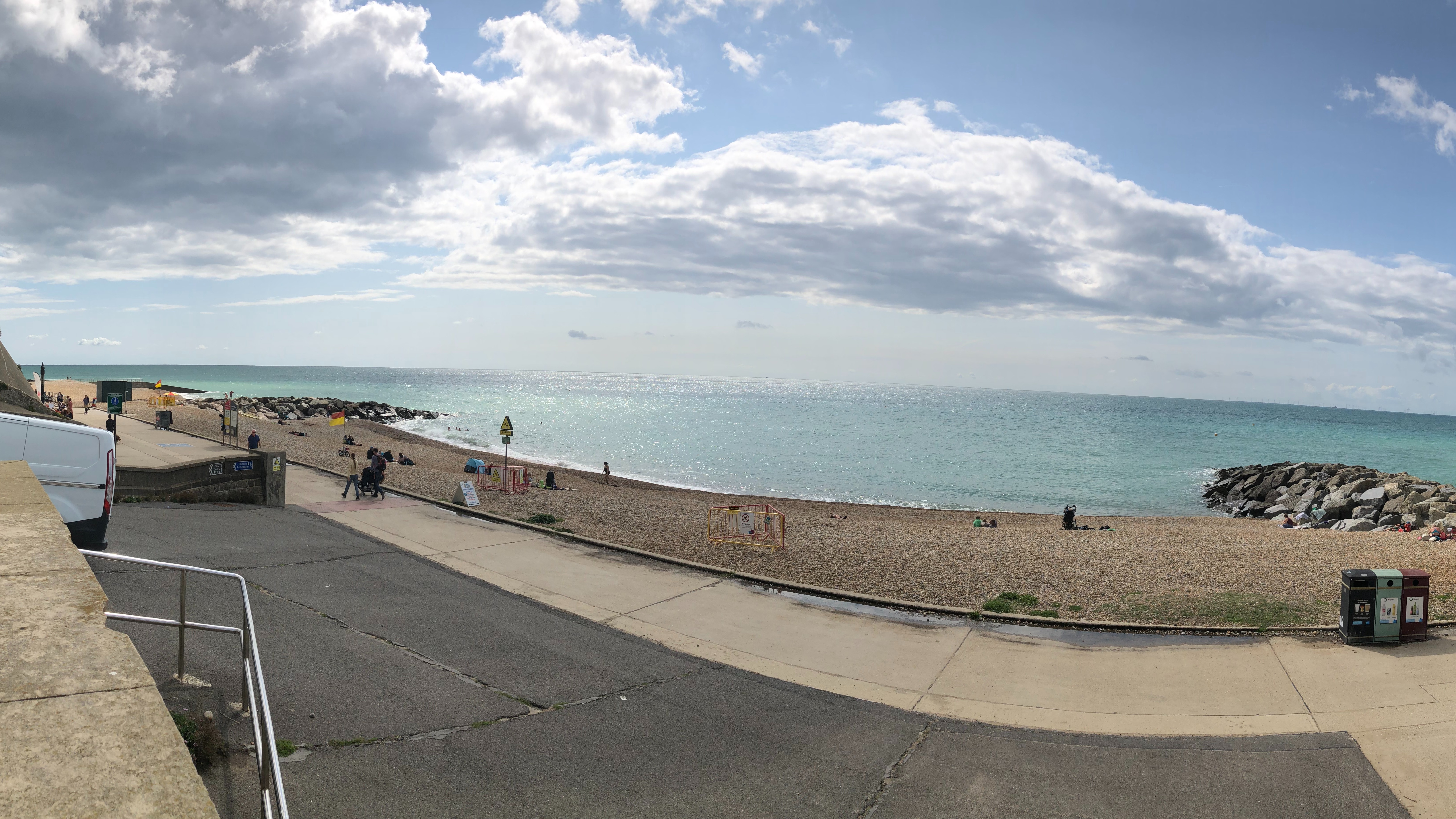 A horizontal landscape image of the beach at Rottingdean. It is a sunny day - the sky is blue with scattered white clouds. In the distance people can be seen enjoying the sun.