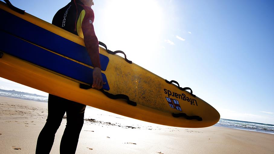 A Lifeguard stands facing the sea on a sunny day. They are holding a Lifeguards branded surfboard. 