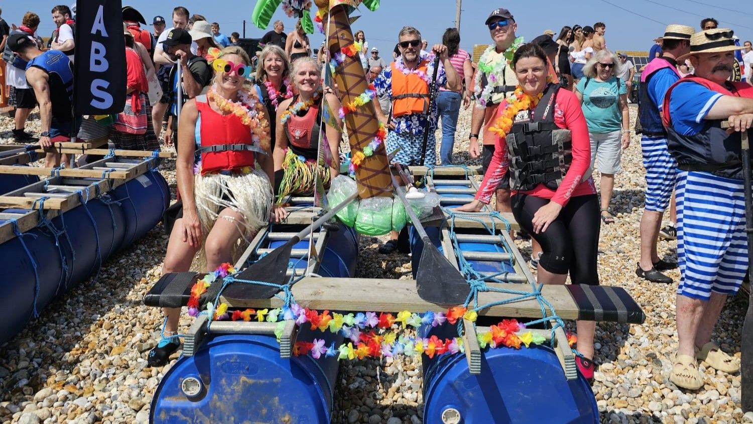 Group in colourful fancy dress posing with a decorated homemade raft on a pebble beach, with a crowd behind.