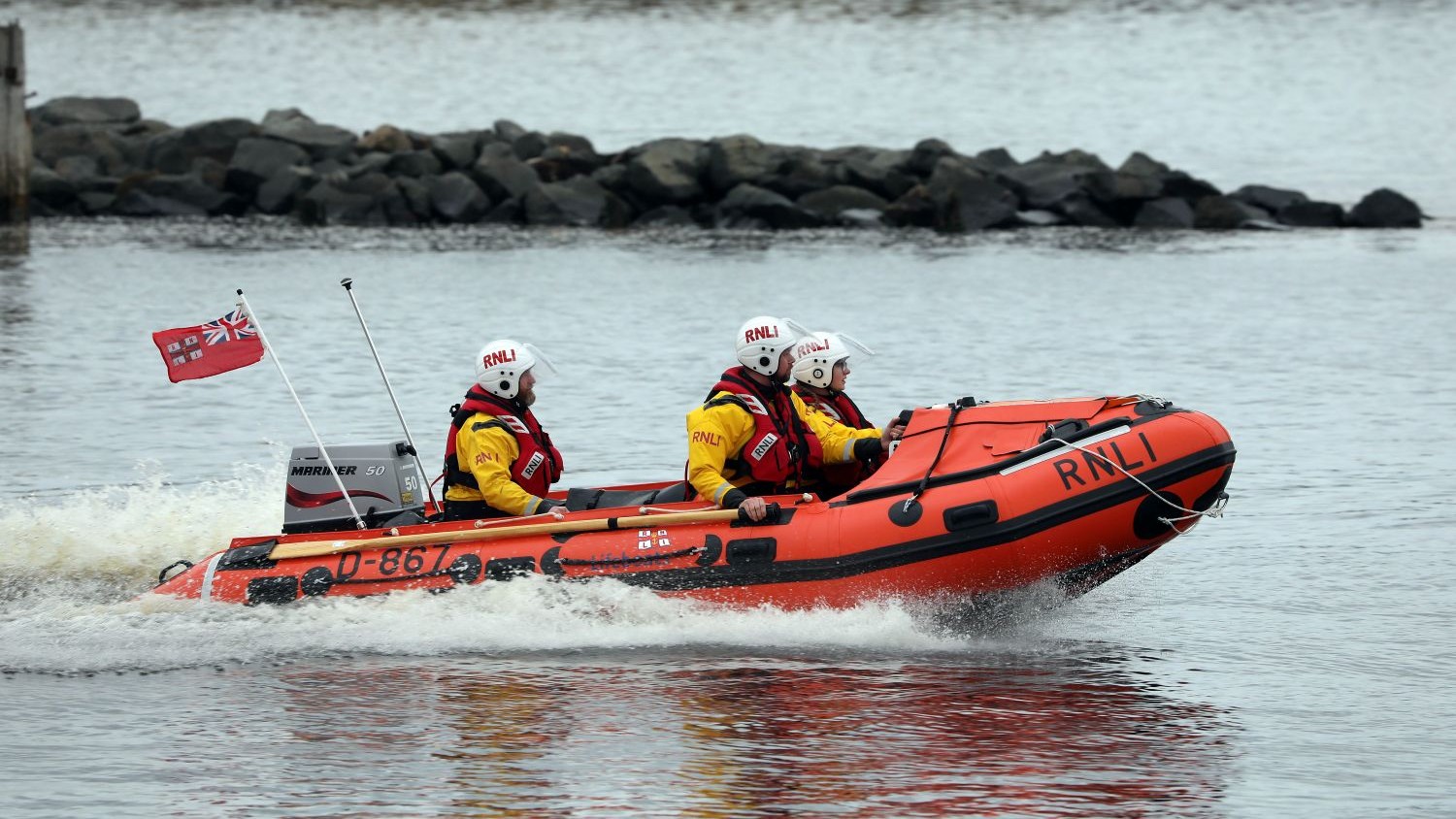 A small rib with three crew on grey water going past a rocky sea wall.