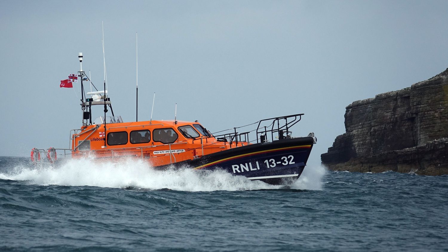 RNLI lifeboat marked RNLI 13-32 speeding through choppy water near rocky cliffs.