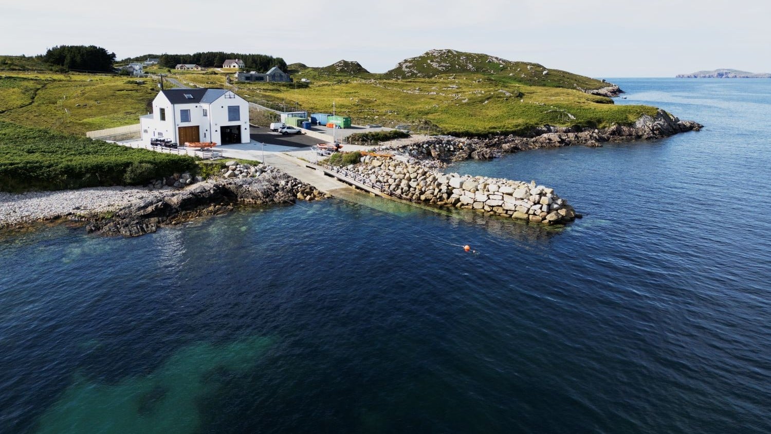 A two-storey white building with slipway and rock sea wall on a grassy and rocky coast.
